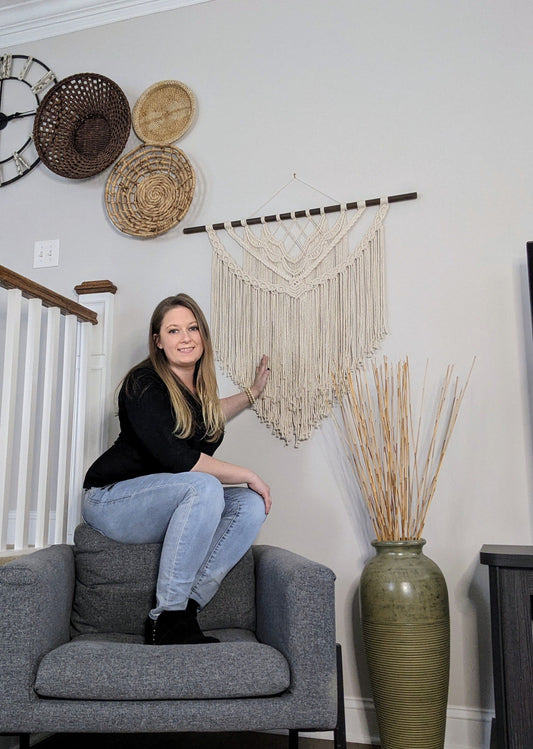 Cassie, the shop owner is perched on the back of a grey arm chair. One arm is outstretched and touching her handmade macrame wall tapestry. She is wearing a black sweater, jeans and black boots. 
